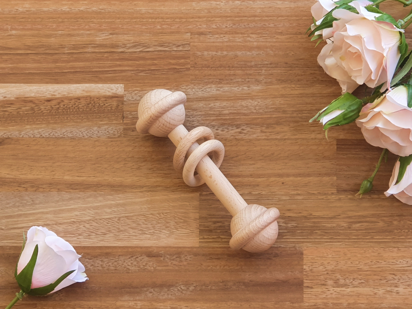 A natural light wood baby teething toy with a spiral ring design, displayed diagonally on a wooden surface with pink roses in the corners.