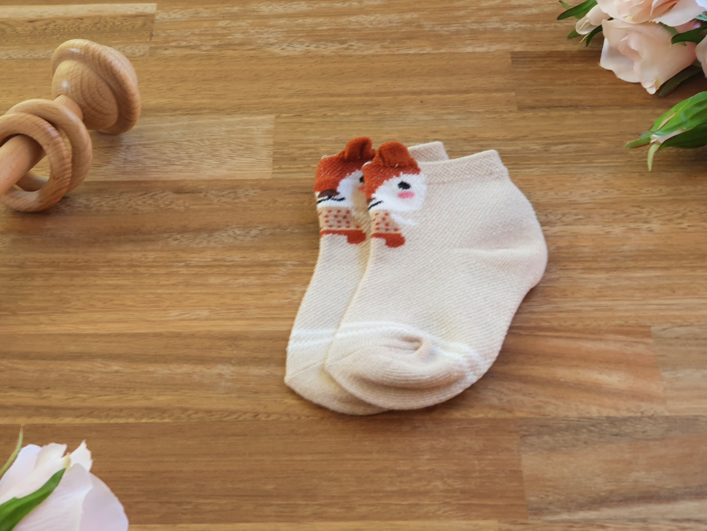 A pair of cream-colored ankle socks with embroidered red and white fox faces on the toes, displayed on a wooden surface with decorative props.