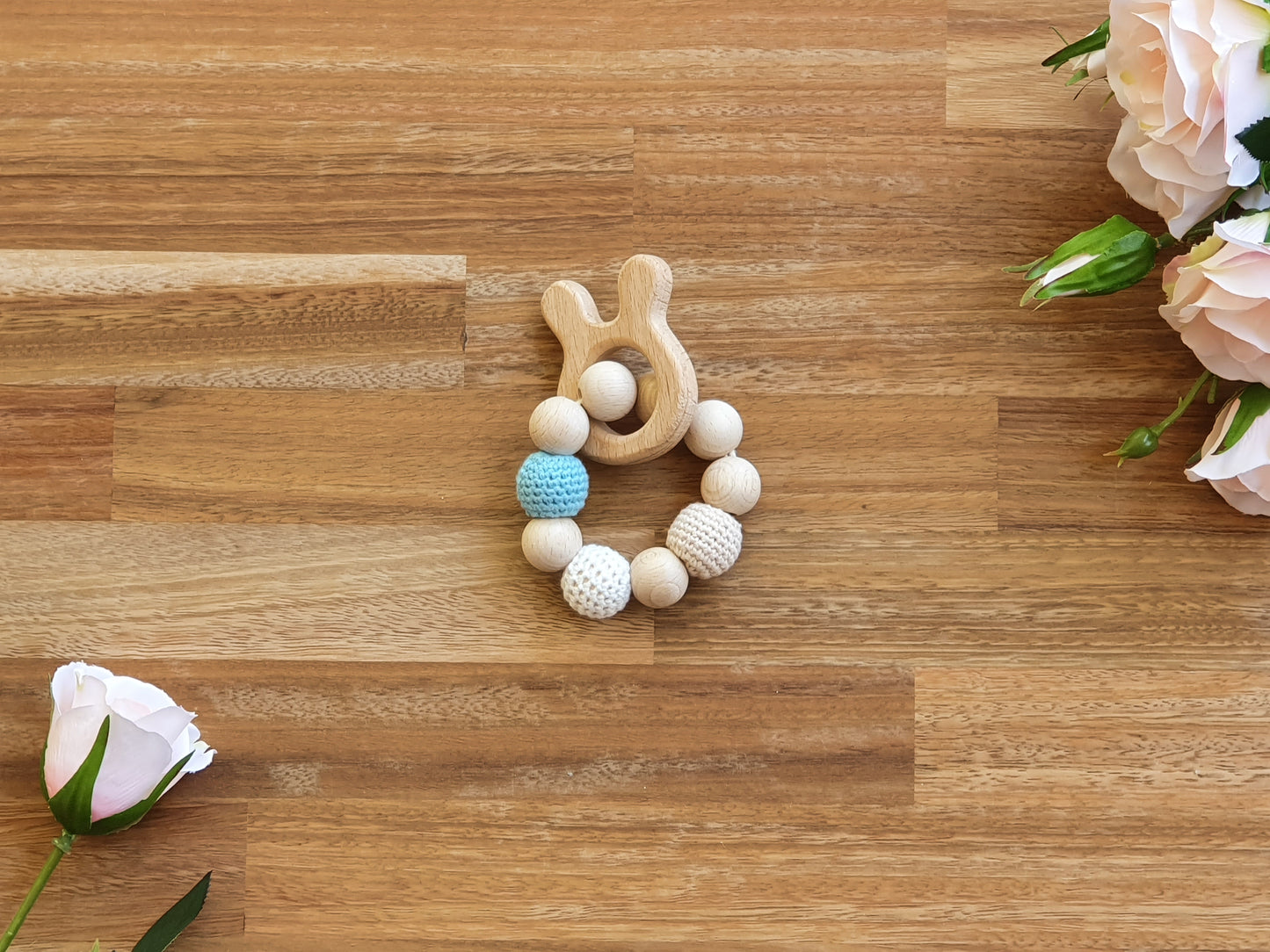 A wooden baby teether with a bunny silhouette and a ring of natural wood and textured beads, including one light blue bead, displayed on a wooden surface with pink roses.