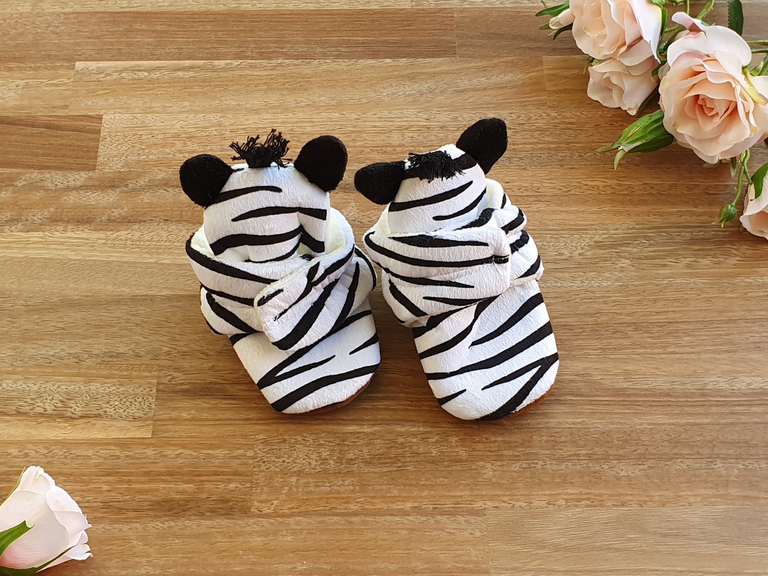 A pair of baby bootie slippers designed to look like zebra heads, featuring black and white stripes with black ears, placed on a wooden surface with decorative pink roses.