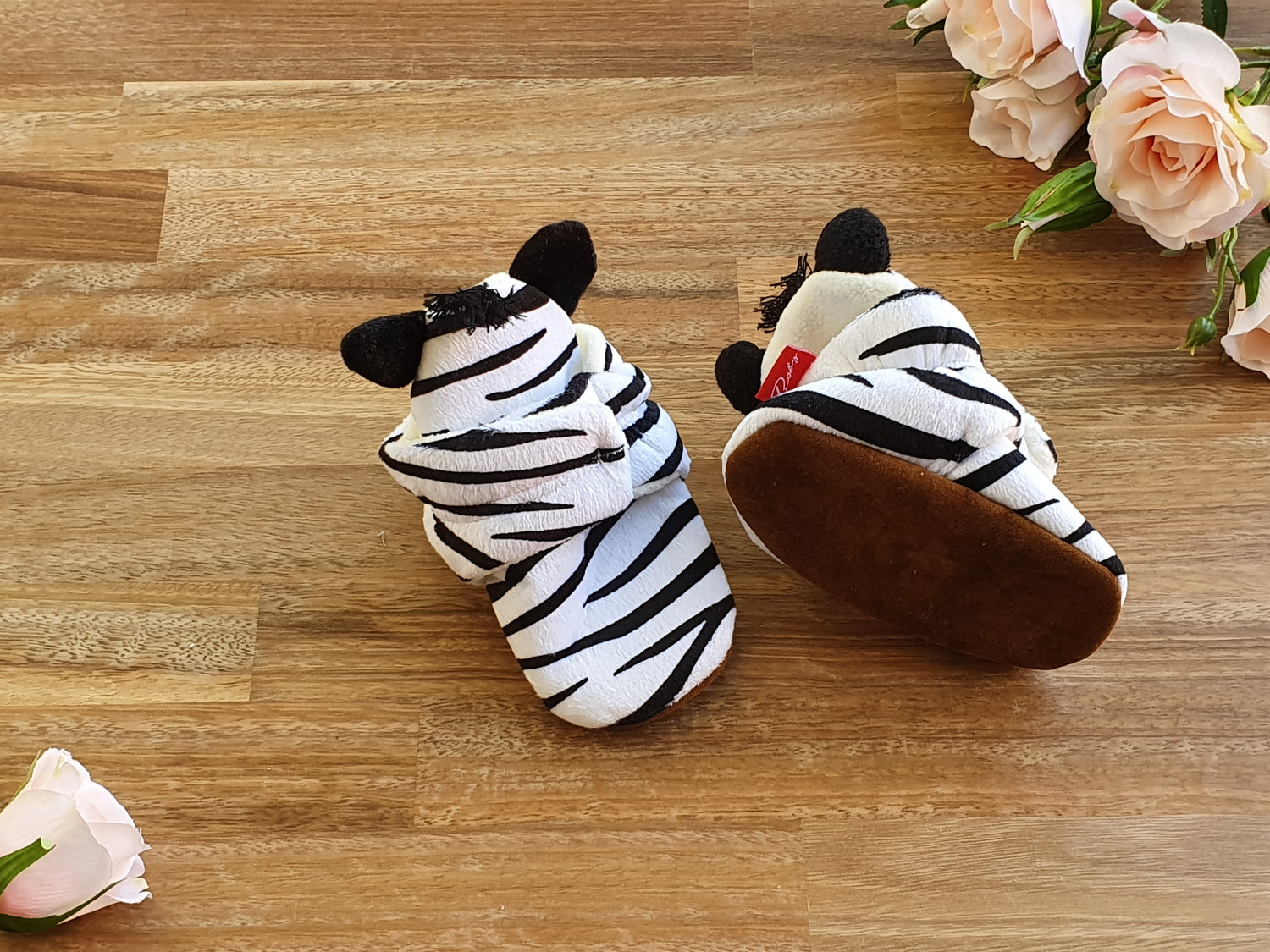A pair of soft baby booties with a zebra print pattern, featuring black plush ears and a brown sole, displayed on a wooden surface with decorative pink roses.