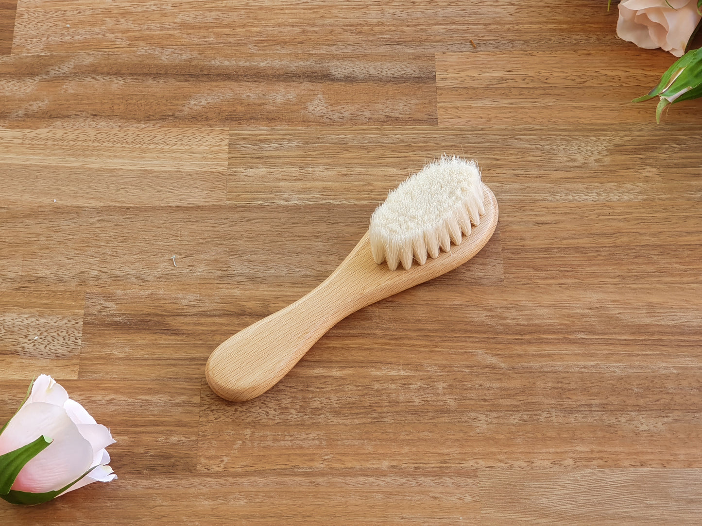A small wooden hairbrush with off-white bristles, placed diagonally on a brown wood-grain surface with decorative pink flowers in the corners.