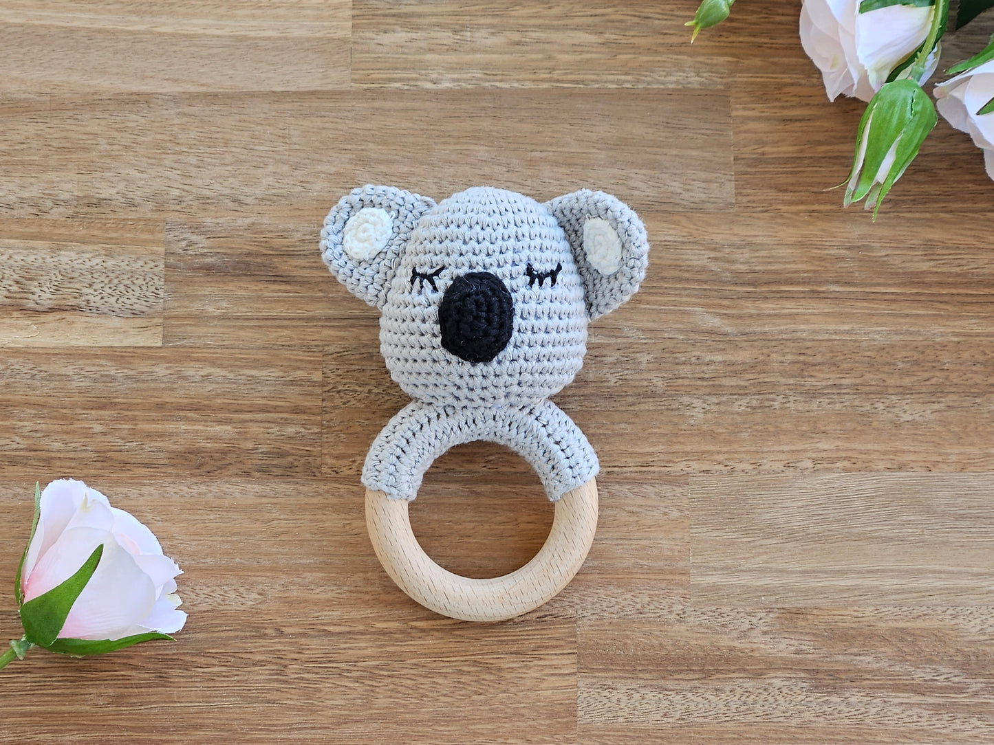 A crocheted koala baby teether rattle with a light gray knit body, black nose and closed-eye details, and a natural wooden ring handle.