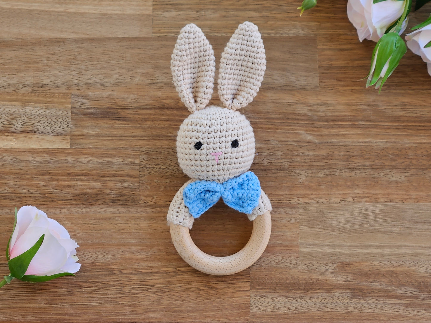 A crocheted bunny teething ring toy with long cream-colored ears, a blue bow tie, and a natural wooden ring, displayed on a wooden tabletop with decorative pink flowers.