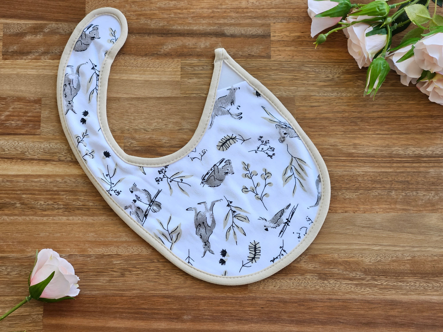 A white fabric baby bib with a beige border, featuring a delicate print of birds, branches, and leaves in grey and olive green.