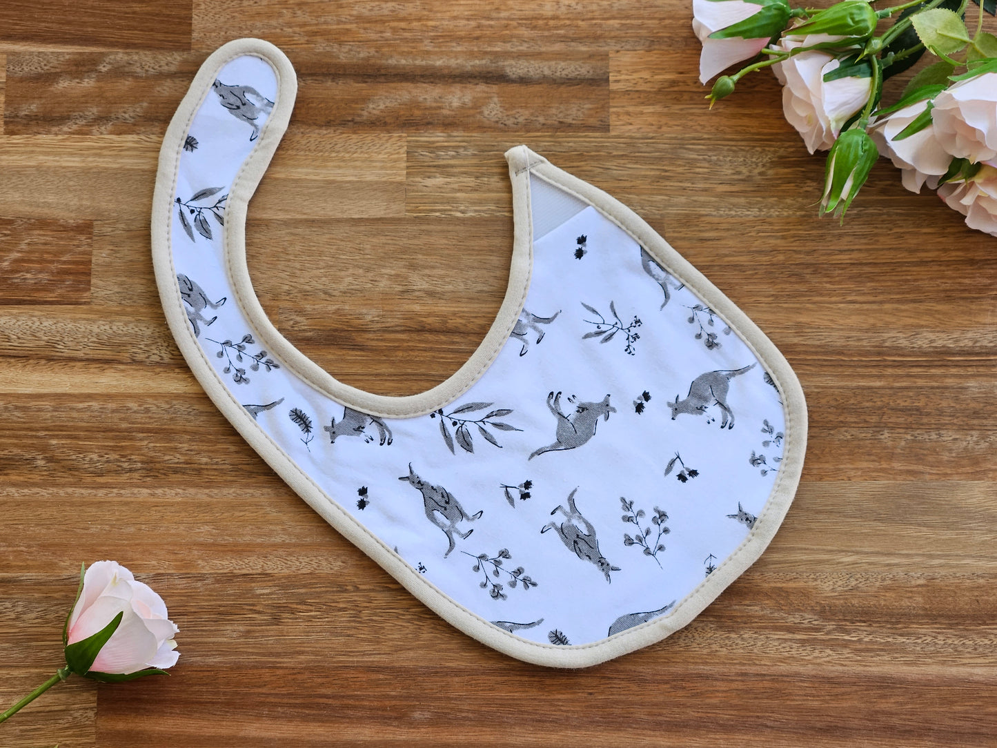 A baby bib on white cotton, printed with grey kangaroo and leaves, and finished with a beige border.