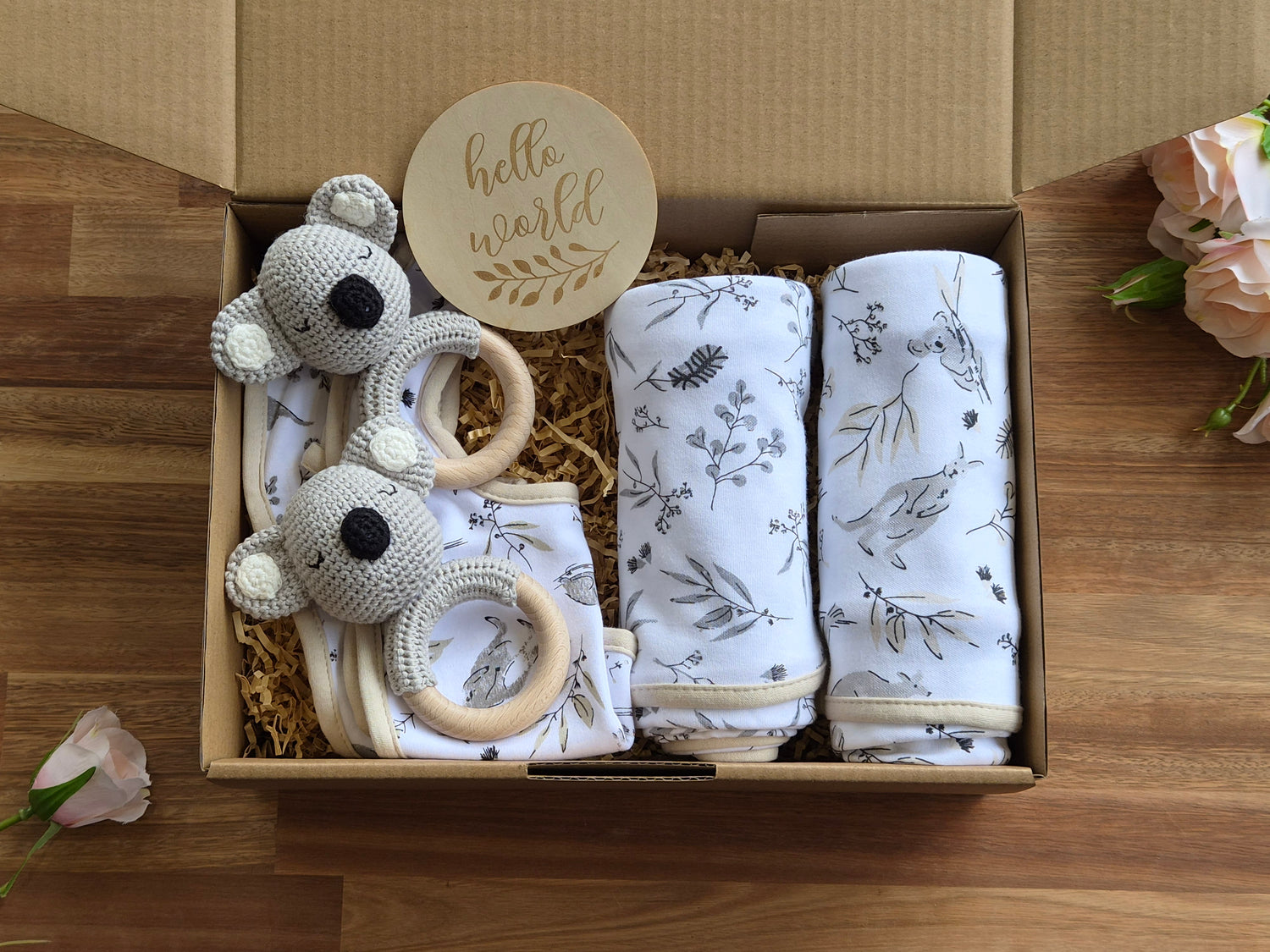 A baby gift box, featuring two white muslin swaddle blankets with grey and gold Aussie print, a grey crocheted koala teether with a wooden ring, and a round wooden tag that reads 'hello world'.
