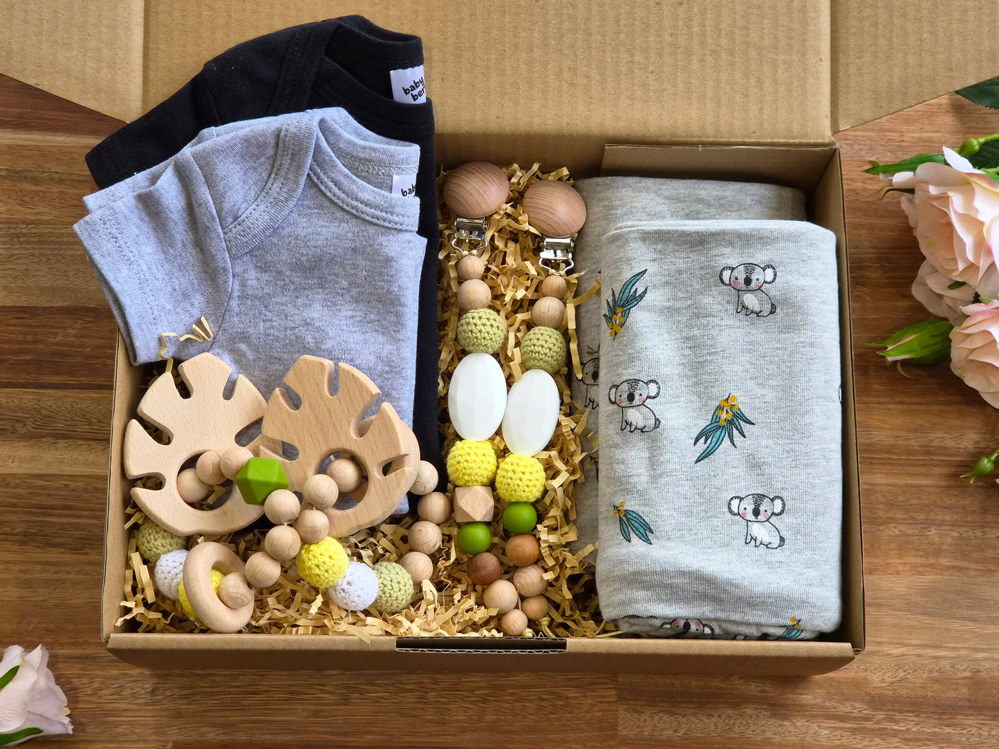 A baby gift box filled with baby items, including a light grey short-sleeve romper, a black short sleeve romper, wooden teething rings, 2 beaded baby pacifiers chains and 2 light grey koala-print jersey cotton blankets.