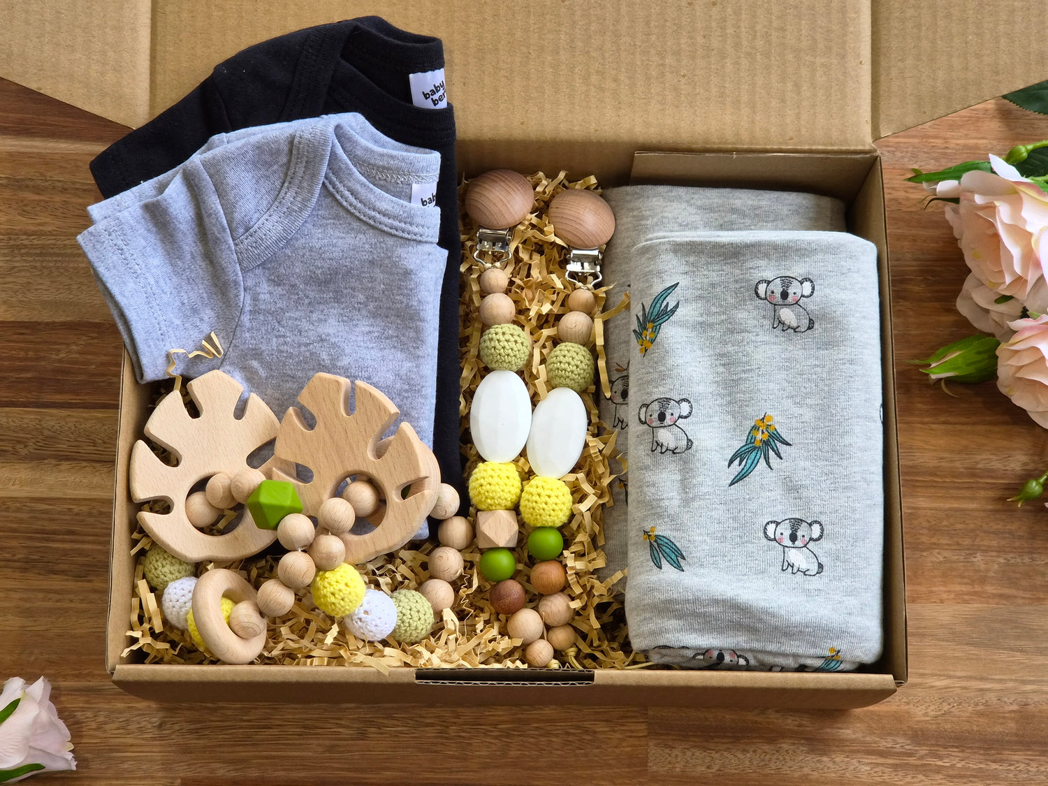 A baby gift box filled with baby items, including a light grey short-sleeve romper, a black short sleeve romper, wooden teething rings, 2 beaded baby pacifiers chains and 2 light grey koala-print jersey cotton blankets.