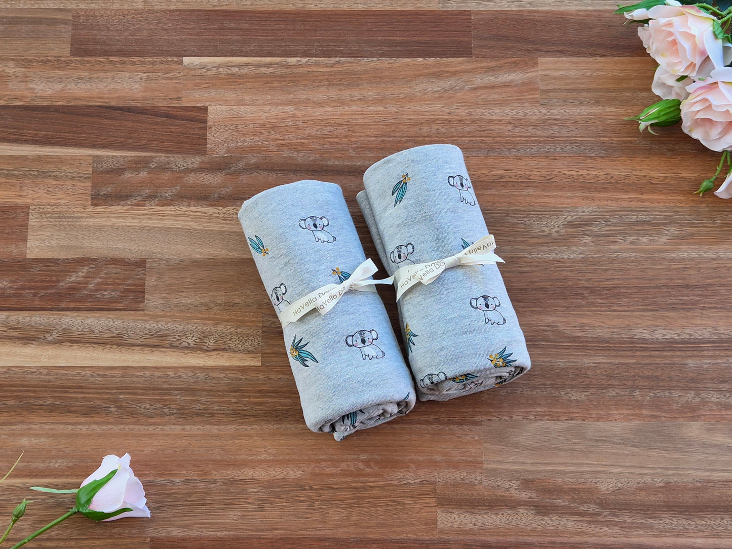 Two rolled baby jersey cotton blankets in a light grey colour with a printed pattern of Koalas and leaves, tied with cream ribbon.