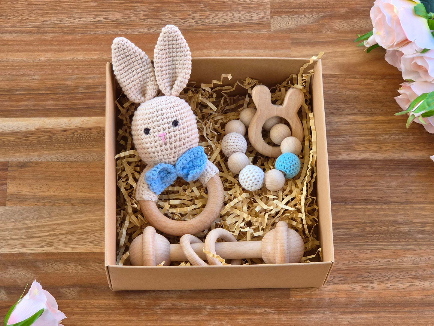 A baby gift set in a brown cardboard box filled with shredded paper, featuring a crocheted bunny rattle with a blue bow, wooden teething toy bar and beaded wooden teether ring displayed on a wooden tabletop with pink flowers in the corners.