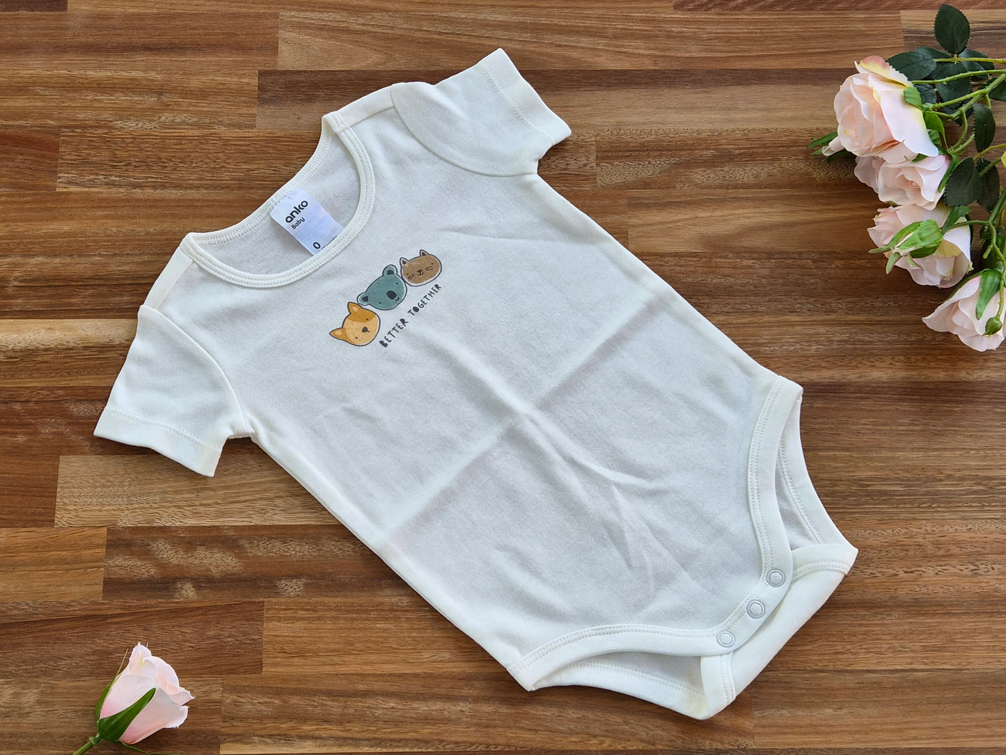 A white short-sleeve baby bodysuit with a small cartoon animal graphic and the text 'little bear' on the chest, laid flat on a wooden surface with pink roses in the corners.