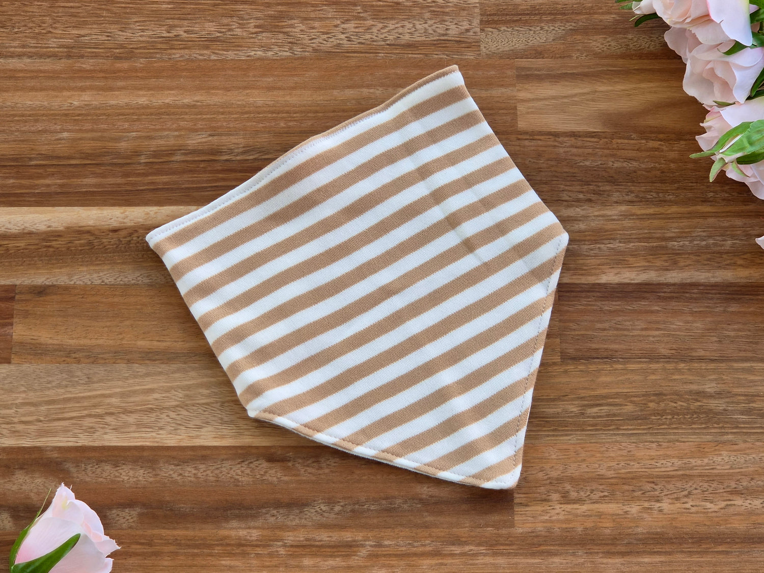 A square bandana with horizontal tan and white stripes, laid flat on a brown wooden surface with pink flowers in the corners.
