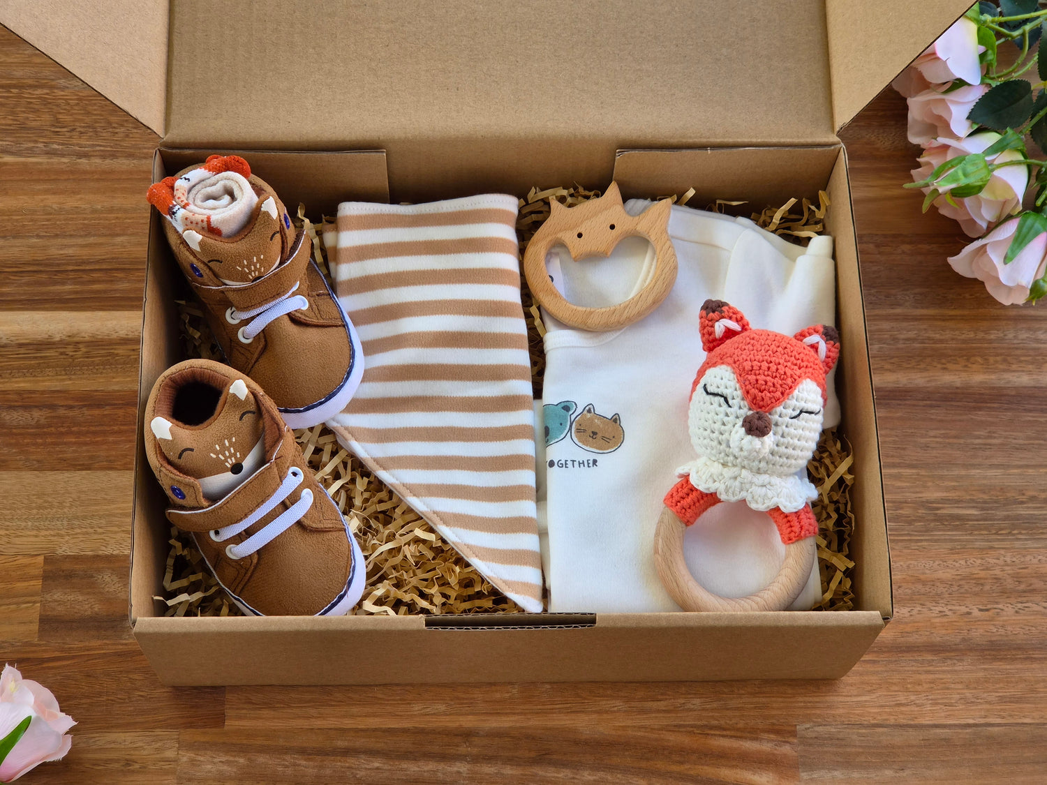 A baby gift set arranged inside an open cardboard box on a wooden surface, including brown baby shoes with a fox face, a striped bandana bib, a white romper with a small bear graphic, a wooden fox teething toy, and a crocheted fox rattle.