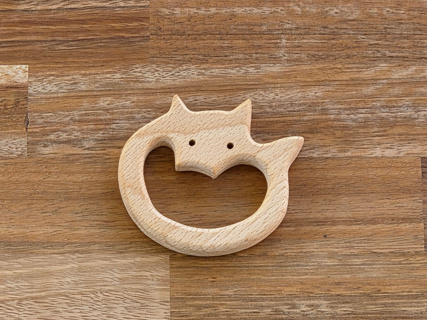 A light natural wood fox-shaped teething ring with a carved face and small eye holes, displayed on a brown wood-grain surface.