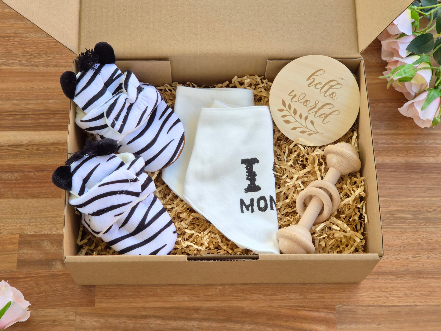 A baby gift box filled with a zebra-striped plush slipper, a white bandana with 'I'M love MON and I love DAD' text, a wooden baby teething ring, and a round wooden 'hello world' sign, arranged on a wooden surface.