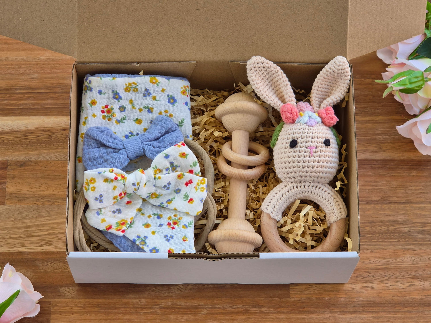 A baby gift set arranged inside an open cardboard box on a wooden surface, featuring a floral bandana bib, 2 matching bow headbands, wooden teething bar toy, and a crocheted bunny teether with pink flower headband.