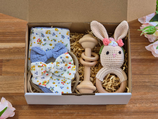 A baby gift set arranged inside an open cardboard box on a wooden surface, featuring a floral bandana bib, 2 matching bow headbands, wooden teething bar toy, and a crocheted bunny teether with pink flower headband.