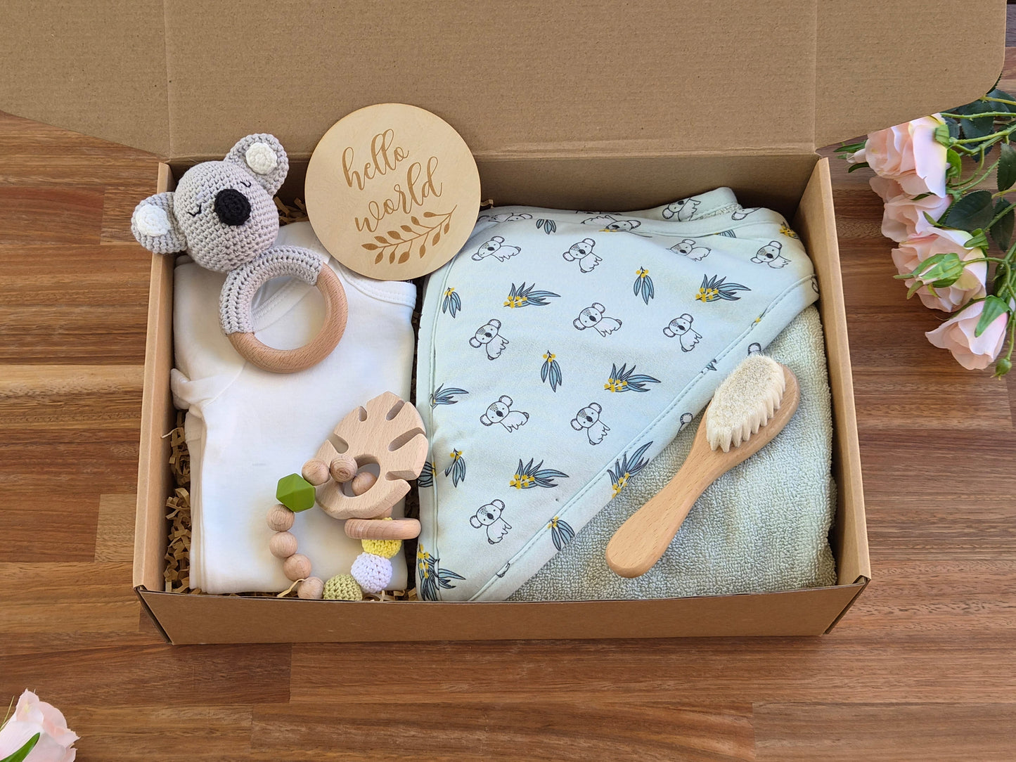 A baby gift box set on a wooden table, containing a white baby onesie, a light green patterned hooded towel, a wooden teething ring, a wooden hand-shaped teething toy, a plush koala rattle, and a wooden baby brush. A round wooden disc that reads 'hello world' is also included.