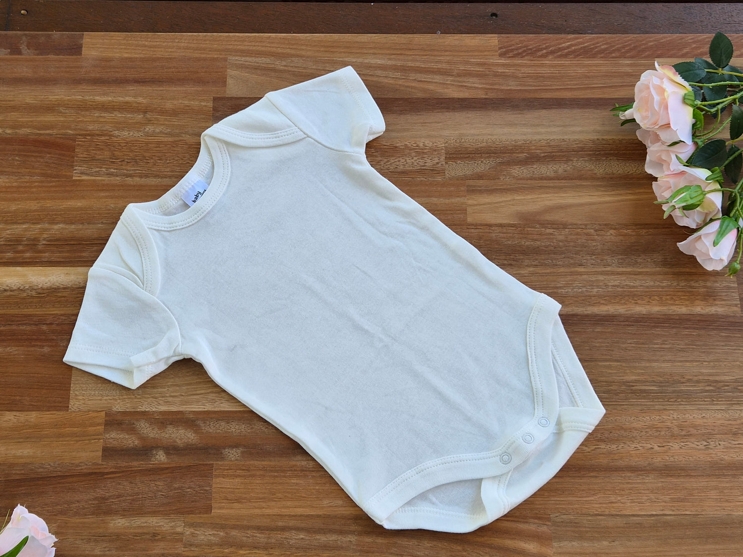 A plain white short-sleeve baby bodysuit laid flat on a wooden surface, with pink roses placed near the top right corner.