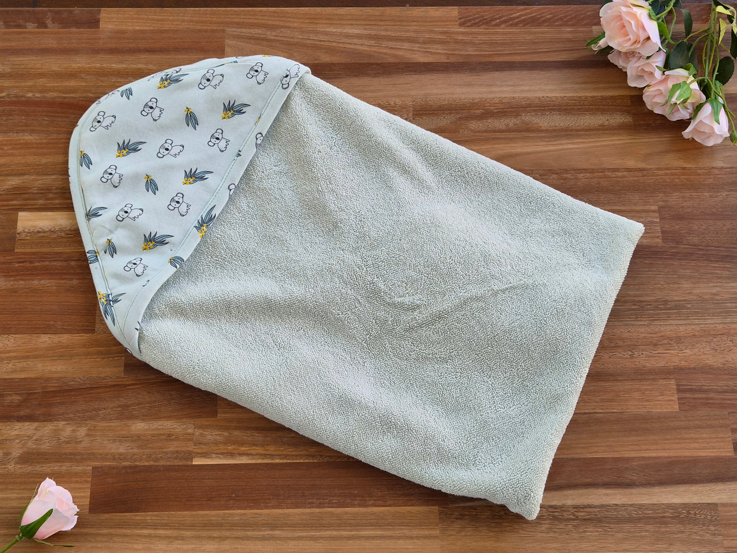 A baby hooded towel laid flat on a wooden surface, with a soft pale sage-green body and a hood lined in a light green fabric printed with small white koala motifs and tiny yellow accents. Pink flowers are placed around the edges as styling props.