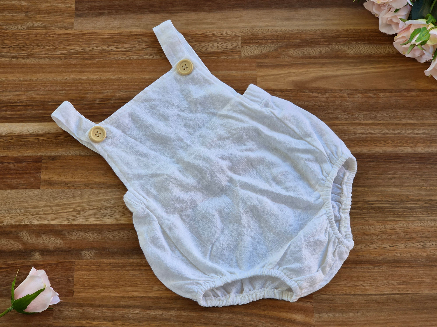 A white cotton baby romper with wooden buttons and shoulder straps, laid flat on a wooden surface with decorative pink flowers in the corners.