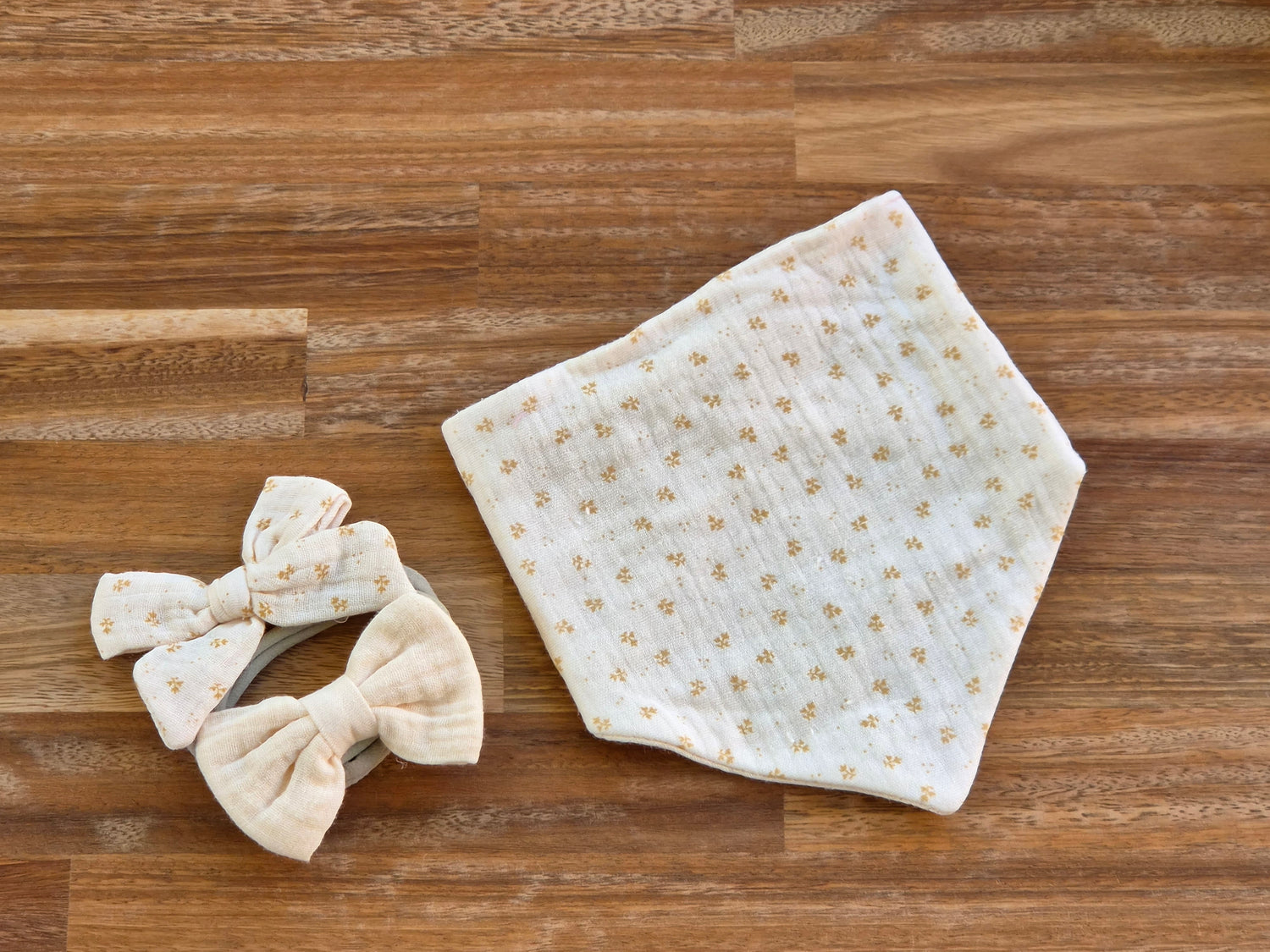 A folded cream-colored muslin cloth bandana bib with a small gold floral pattern is displayed on a brown wooden surface next to a matching baby headband with two fabric bows.