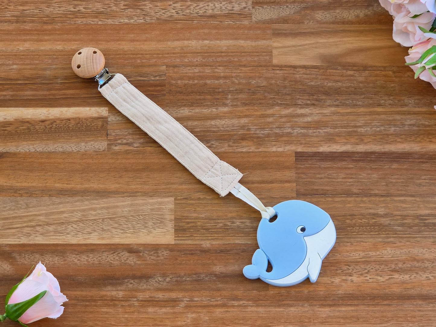 A baby muslin pacifier clip with a light blue whale-shaped silicone teether a beige fabric strap.