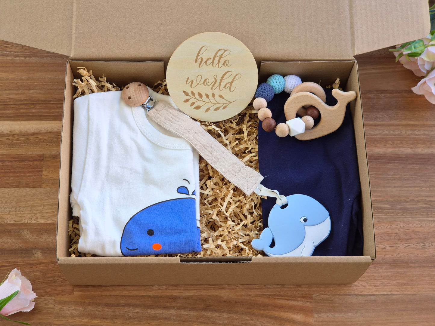 A baby gift set in a cardboard box filled with shredded paper, featuring a white t-shirt with a blue whale graphic, a navy leggings, a wooden sign reading 'hello world', a wooden bead teether ring, and a muslin pacifier with silicone whale.
