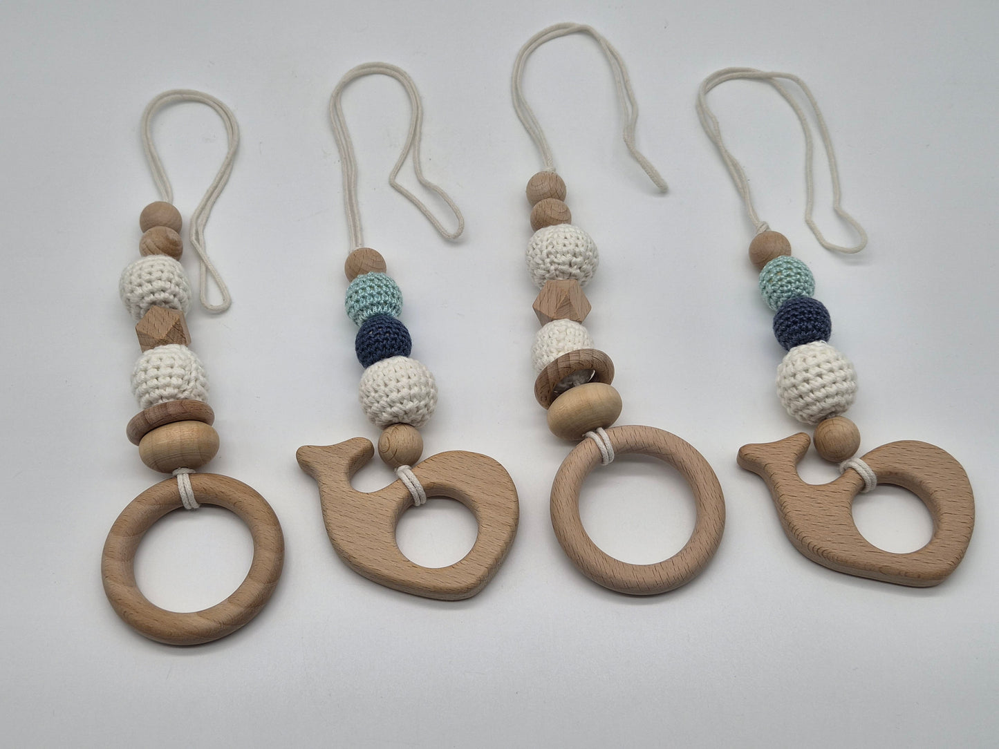 Four wooden baby gym hanging toys with knotted beaded straps, arranged on a wooden tabletop with pink flowers in the corners.