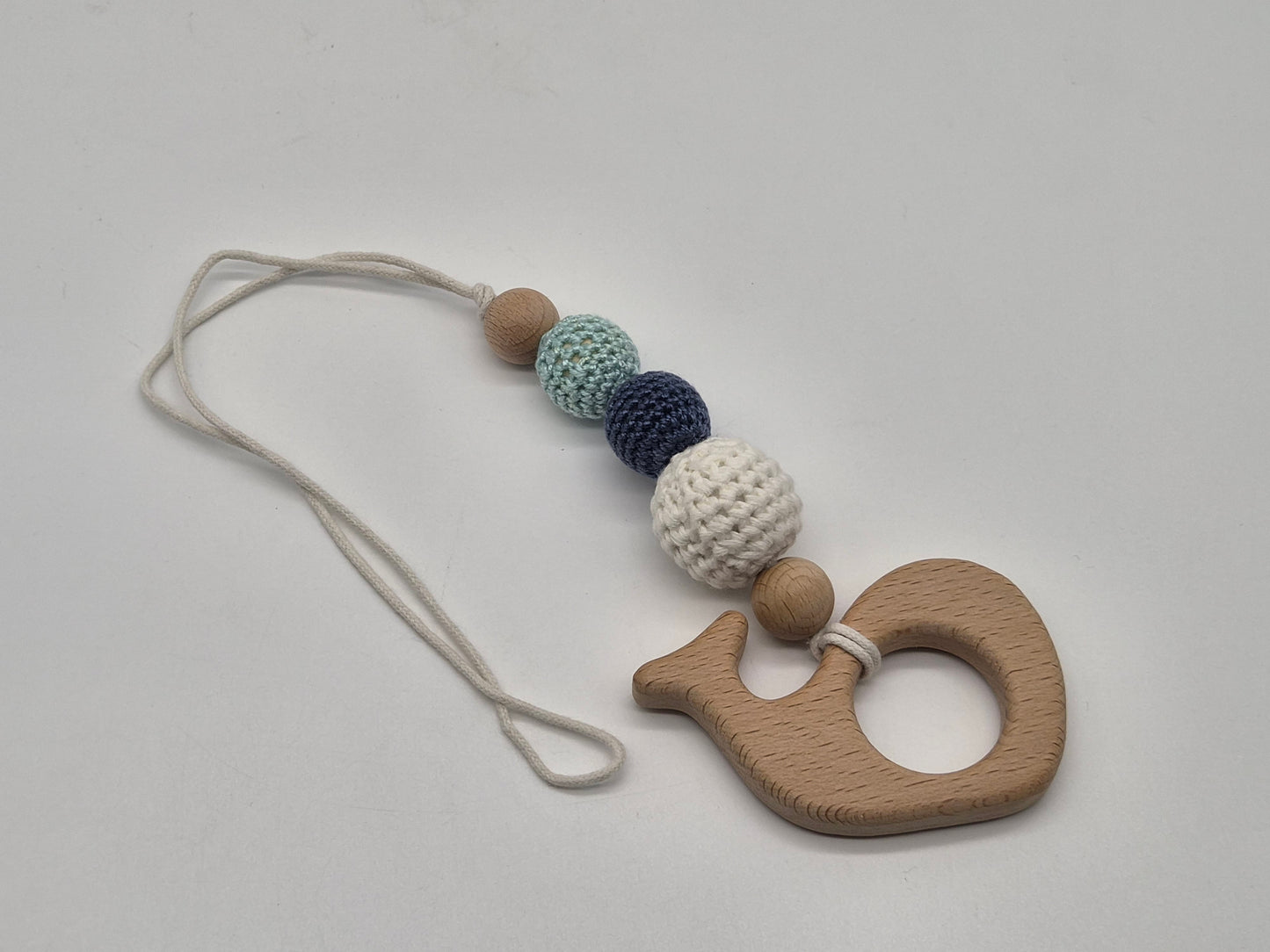 A wooden whale-shaped baby gym toy with a cream-colored string and a series of textured beads in light brown, mint green, navy blue, and cream, displayed on a neutral light gray background.