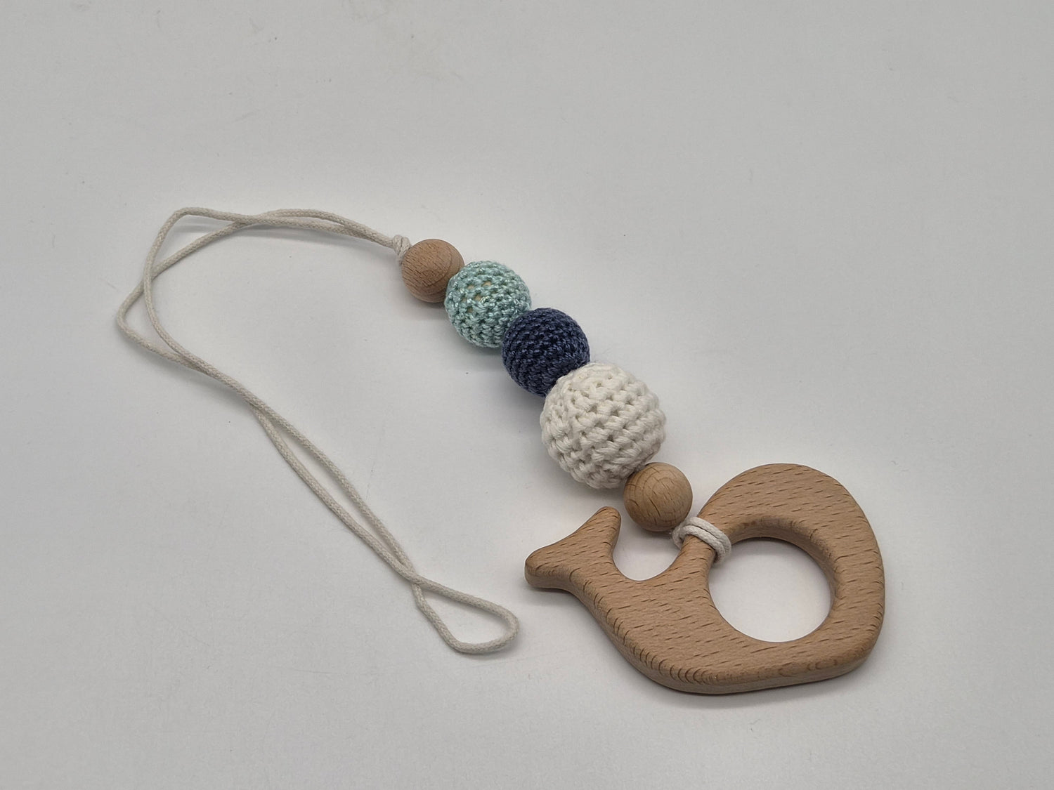 A wooden whale-shaped baby gym toy with a cream-colored string and a series of textured beads in light brown, mint green, navy blue, and cream, displayed on a neutral light gray background.