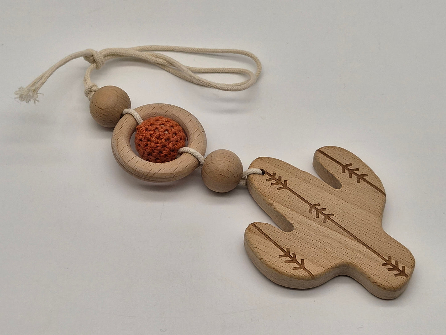 A wooden baby teether and rattle featuring a cactus-shaped wooden piece with engraved leaf patterns, a circular wooden ring, a textured orange crochet ball, and natural wood beads on a cream-colored rope, displayed on a light gray background.