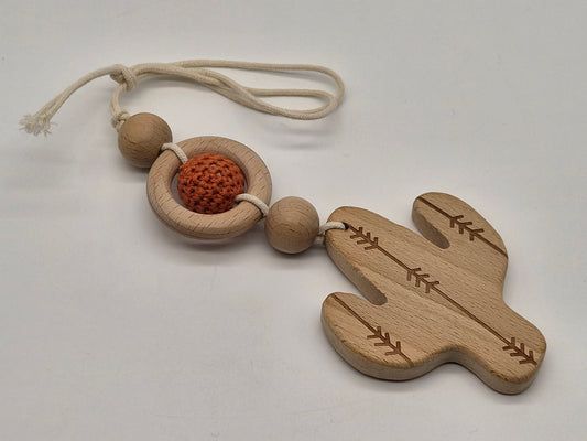 A wooden baby teether and rattle featuring a cactus-shaped wooden piece with engraved leaf patterns, a circular wooden ring, a textured orange crochet ball, and natural wood beads on a cream-colored rope, displayed on a light gray background.
